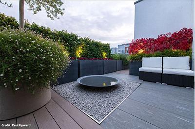 A round water feature in a sculpted garden with a garden furniture sofa adjacent to it and red foliage in planters.